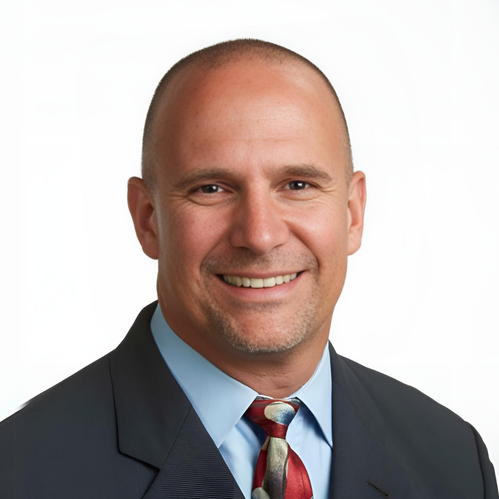 A professional headshot of a smiling man in a suit with a light blue shirt and a patterned tie, against a plain white background.