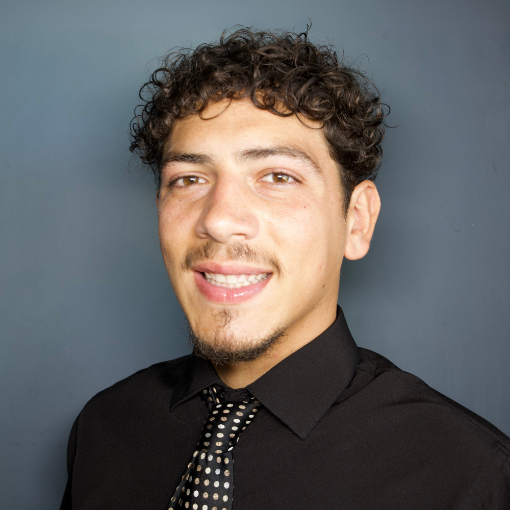 Portrait of a young man with curly brown hair, light facial hair, wearing a black shirt and a black and white polka dot tie, smiling against a solid gray background.