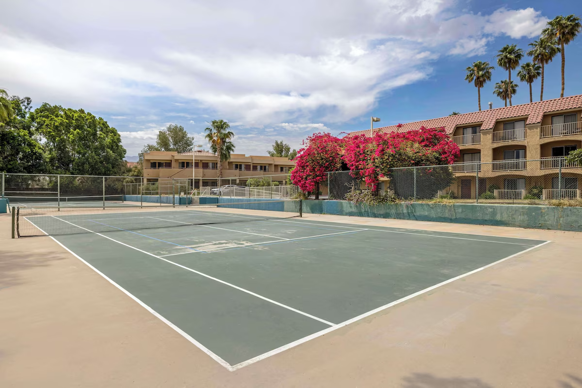 Empty tennis court with net, surrounded by a fence, with colorful flowering bushes and residential buildings in the background under a cloudy sky.