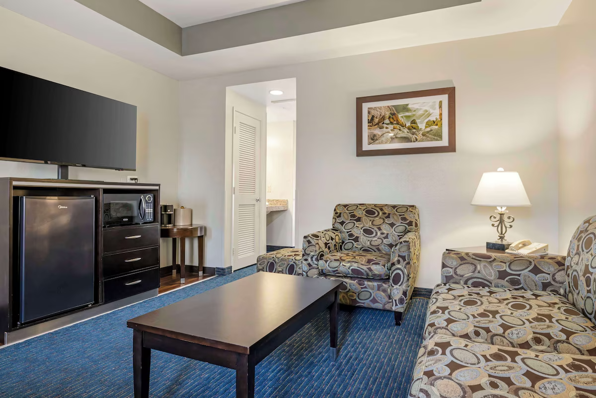 Hotel room with patterned armchair and sofa, coffee table, wall-mounted TV, mini fridge, microwave, small side table with a coffee maker, bed in the background, and a framed photo of a rocky landscape on the wall.