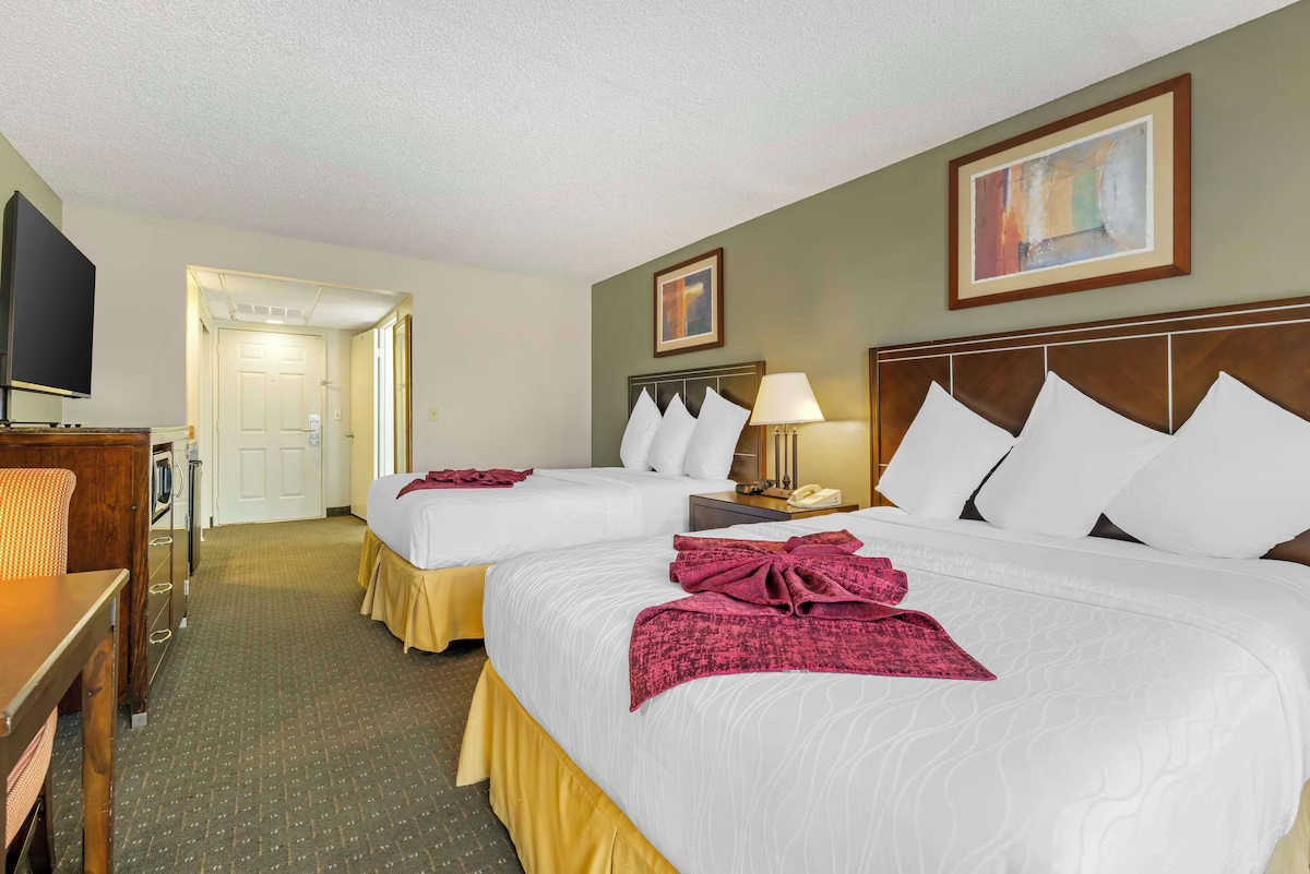 Hotel room with two double beds, each with white linens, pillows, and red folded towels, a nightstand with a lamp, a TV on a wooden cabinet, a chair, and artwork on the walls.