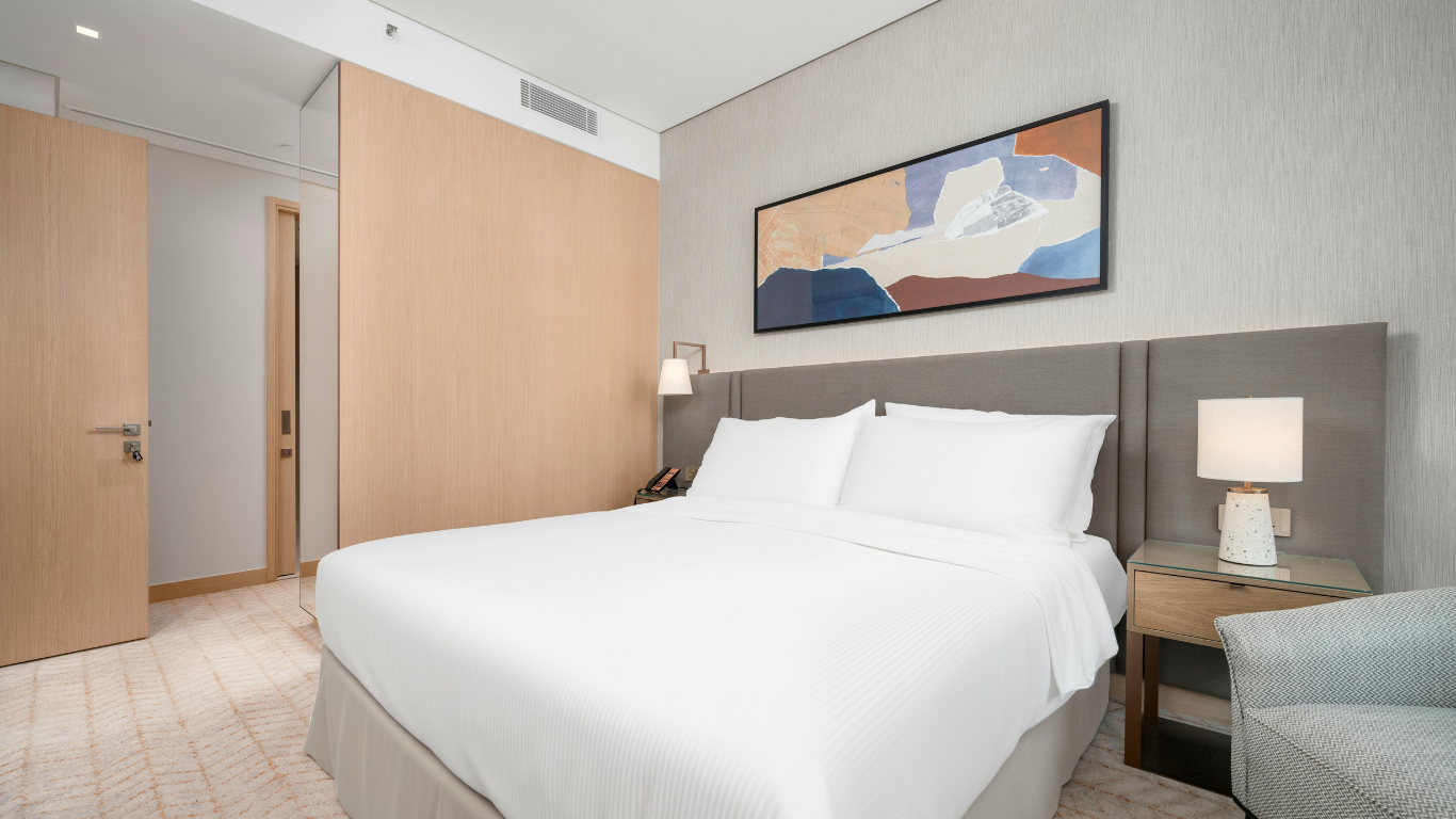 Modern hotel room with a neatly made bed, a bedside lamp, painting above the bed, and neutral decor.