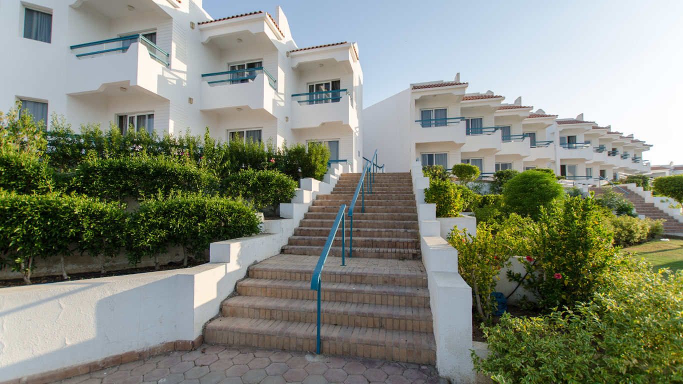 White multi-story buildings with balconies, green bushes, and stairs leading up to the entrances on a sunny day.