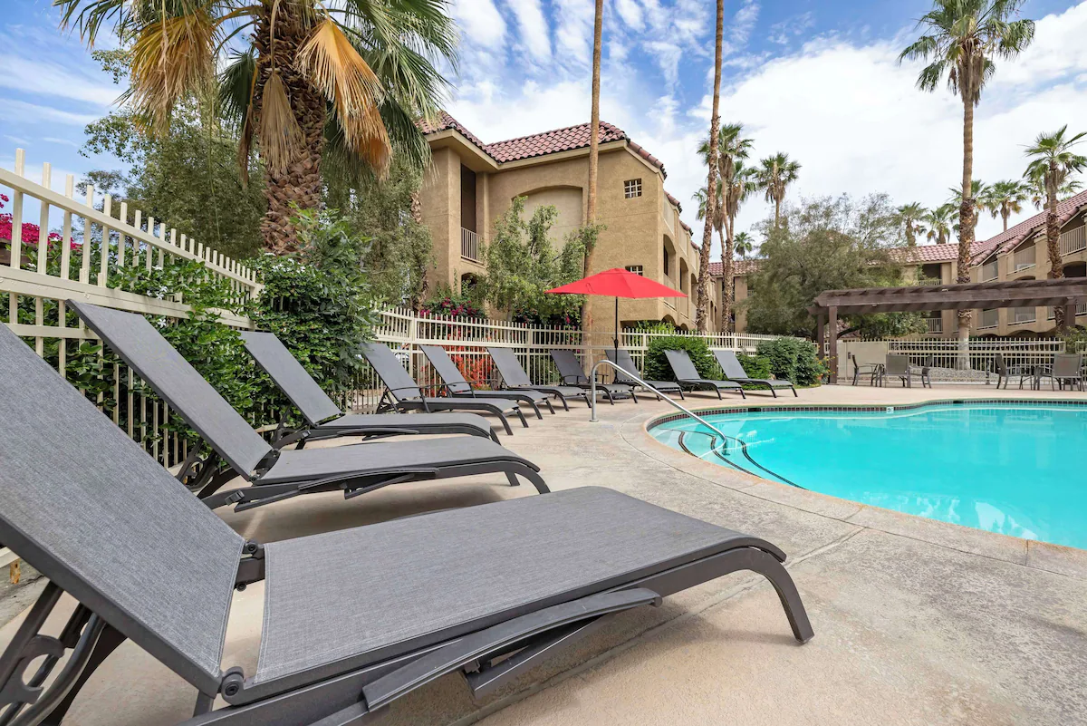 Empty swimming pool surrounded by lounge chairs, umbrellas, and palm trees in a residential complex in sunny weather.