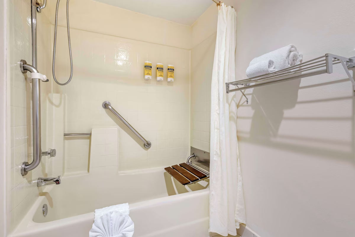 Accessible bathroom with a walk-in shower, grab bars, a foldable seat, white towels on a rack, and a white curtain