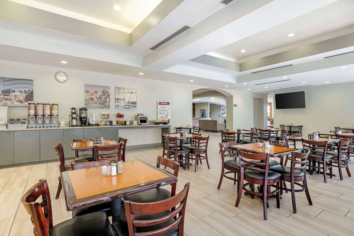 A dining area with multiple wooden tables and chairs, a breakfast buffet station with cereal dispensers and coffee maker, a wall-mounted TV, a clock, and decorative artwork on the walls