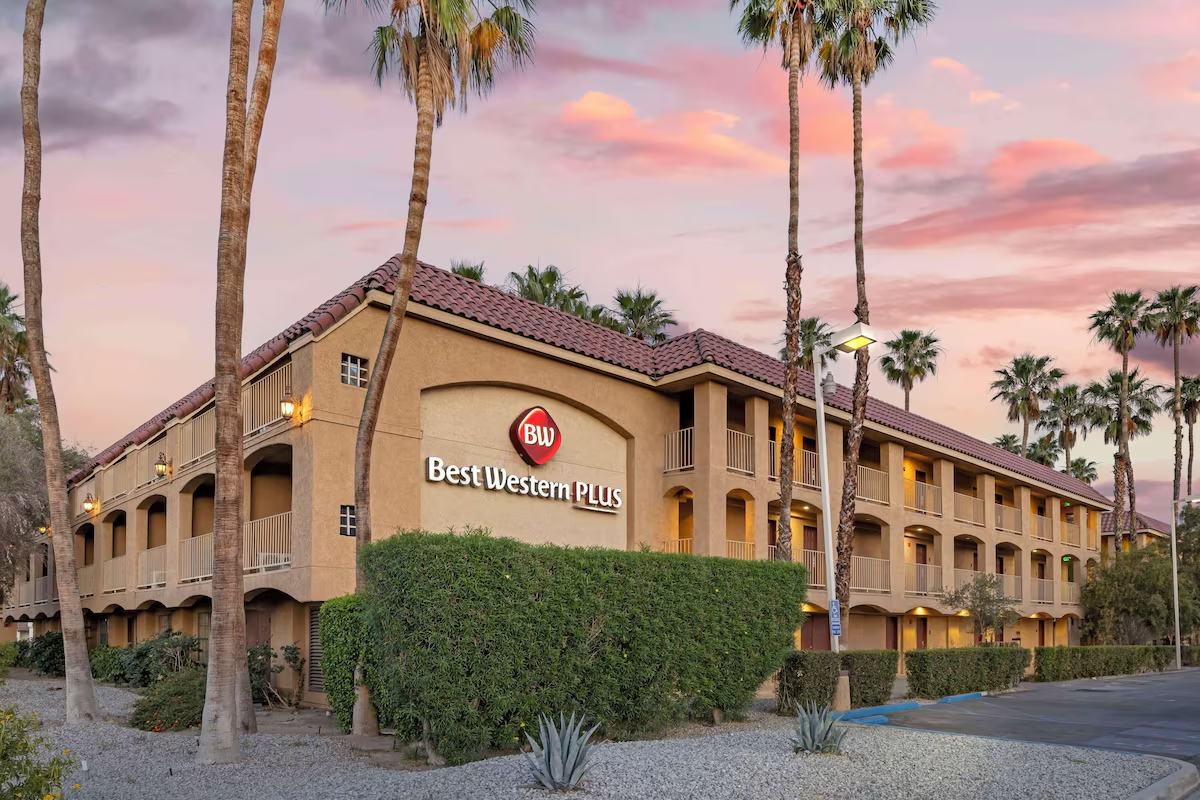 A three-story Best Western Plus hotel with a beige stucco exterior, red tile roof, and multiple balconies, surrounded by tall palm trees during sunset.