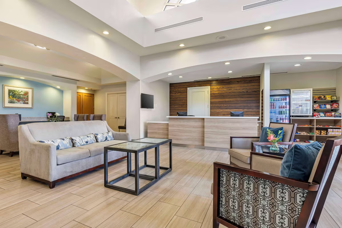 Hotel lobby with beige sofa, armchairs, glass coffee table, reception desk, vending machines, and snack shelves.