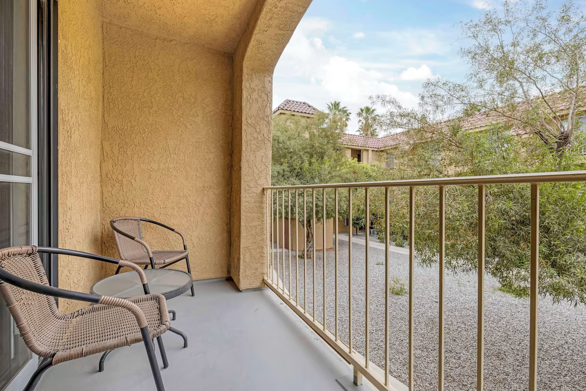 Balcony with two wicker chairs with metal frames and a small round glass-top table, overlooking a gravel courtyard with trees and neighboring buildings in the background.