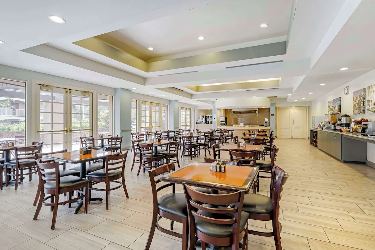 Empty dining area with wooden tables and chairs, large windows letting in natural light, and a breakfast buffet station on the side.