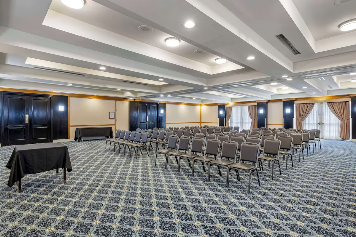 Empty conference room with rows of gray chairs, a black table at the front, patterned carpet, dark wood paneling, large windows with curtains, and ceiling lights.