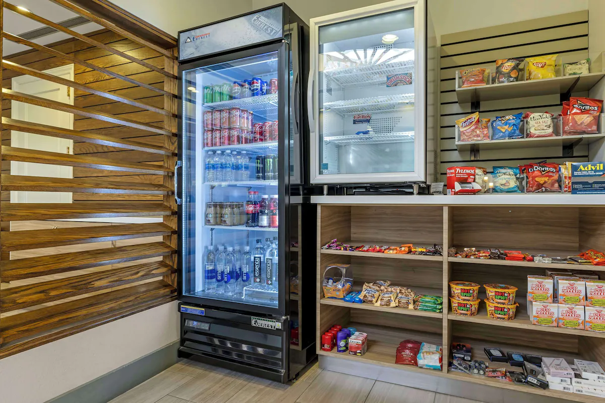 A snack and beverage area with a glass-door refrigerator filled with soda, water, and energy drinks, and wooden shelves stocked with chips, candy, and instant noodles.