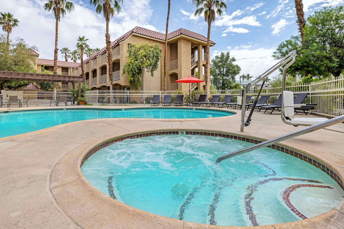 An outdoor swimming pool and hot tub surrounded by lounge chairs and palm trees in a residential complex with beige buildings and a cloudy sky.