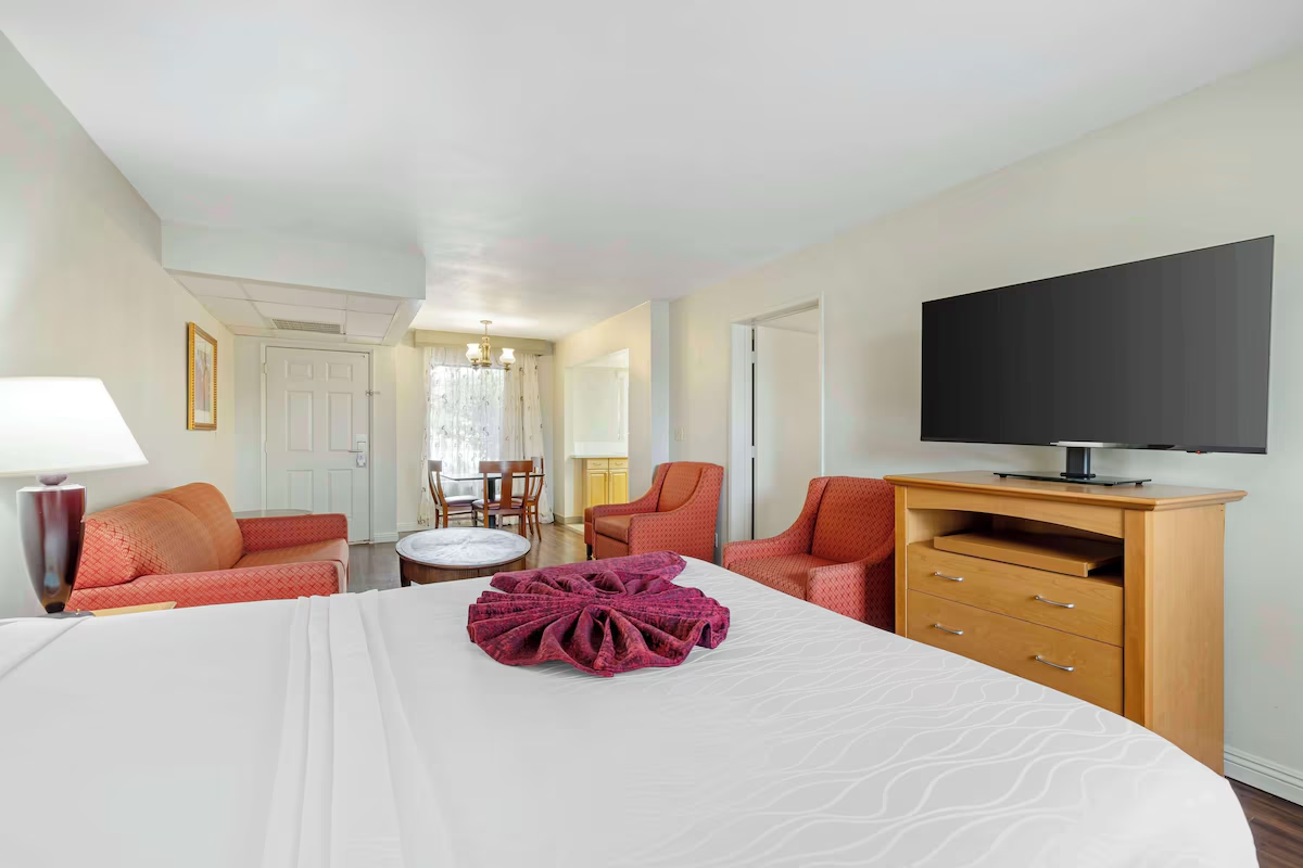 View of a hotel room with a bed, a wooden dresser with a flat-screen TV, red armchairs, a round coffee table, and a dining area with a table and chairs in the background.