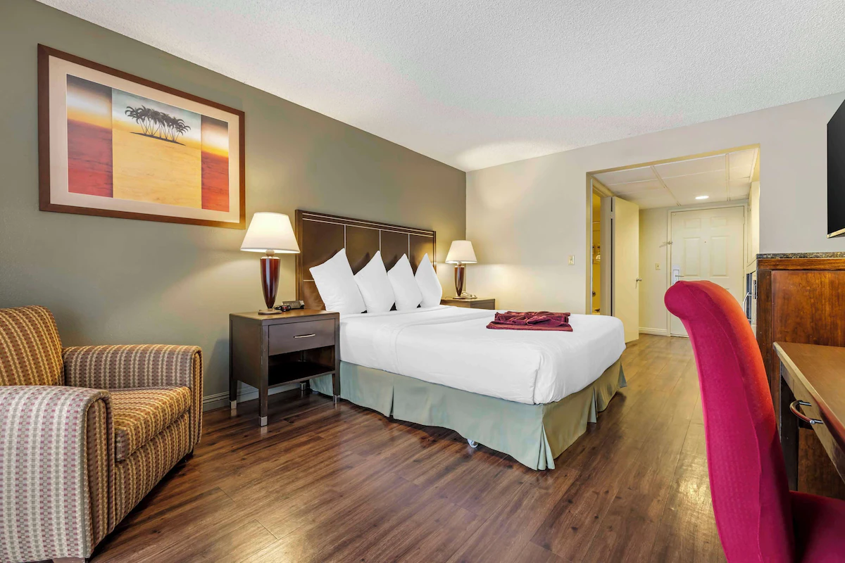 A hotel room with a king-size bed, throw pillows, bedside lamps, a patterned armchair, a red desk chair, and a wooden dresser, with artwork on the wall and hardwood flooring.