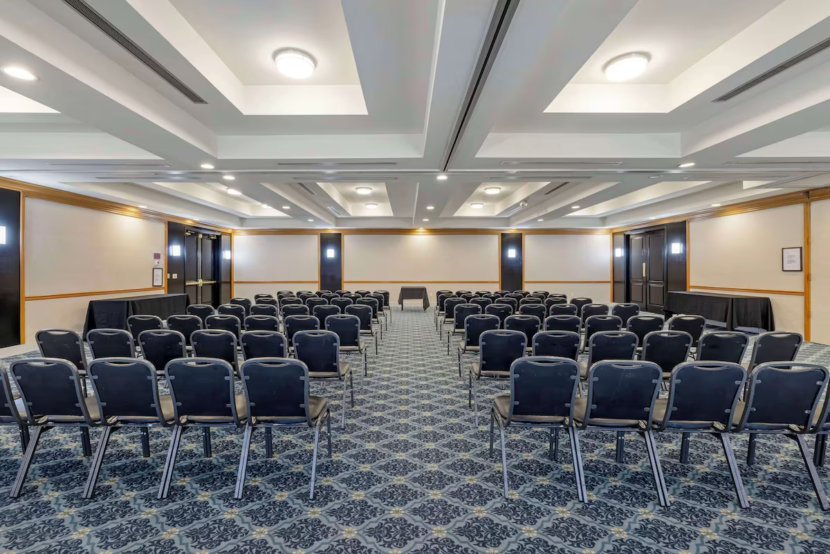 Empty conference room with rows of black chairs on a patterned carpet facing a table at the front, beige walls with wooden trim, and ceiling lights.