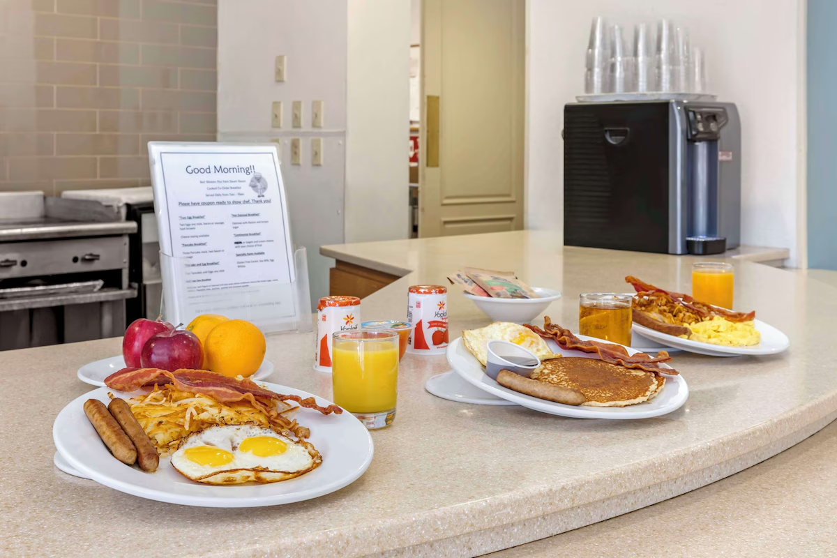 Breakfast served on a counter with two plates featuring eggs, bacon, hash browns, sausage, pancakes, and toast, along with glasses of orange juice and cups of yogurt, apples, and lemons in a cafe setting.