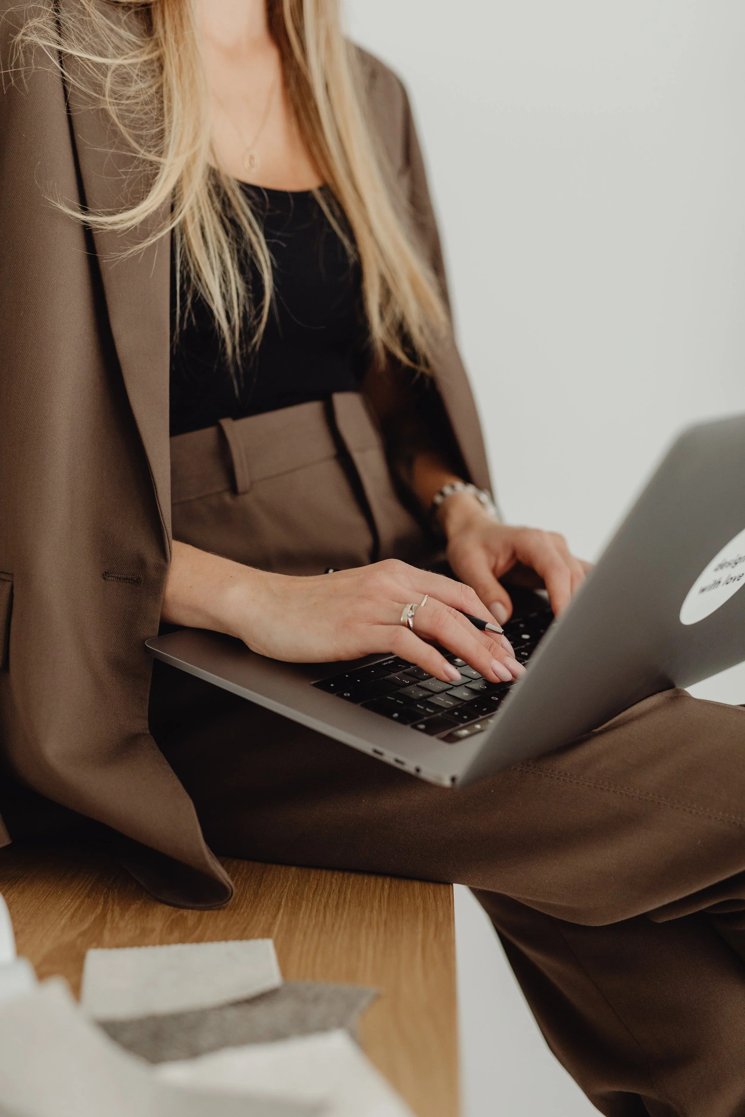 A woman in a brown suit is standing and typing on a laptop placed on a wooden table. She is wearing a black top, a thin necklace, and a ring on her hand. The background is a plain white wall.
