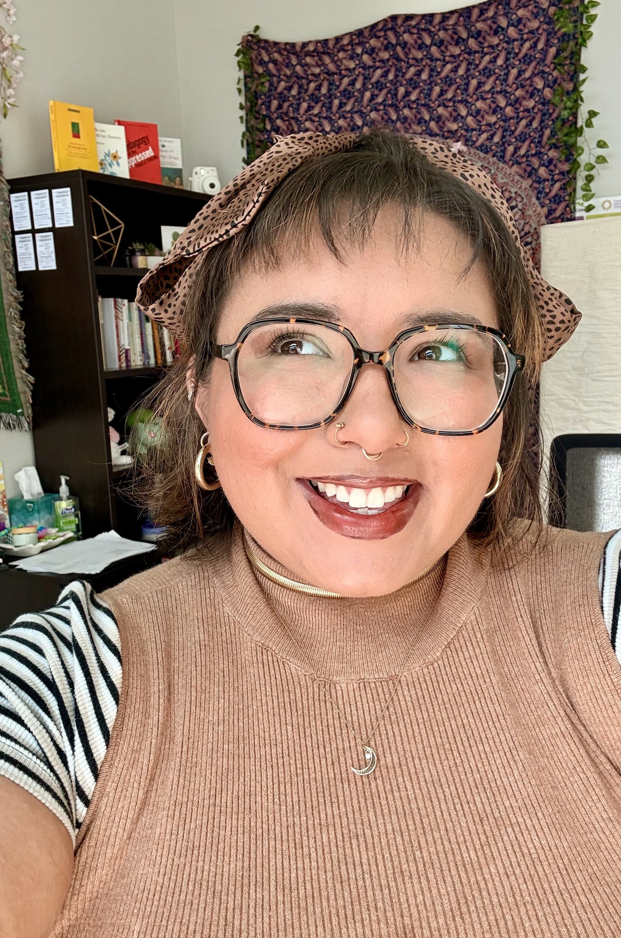 A smiling woman with glasses, a headscarf, gold hoop earrings, and layered necklaces, taking a selfie in a room with books and decorative items in the background.