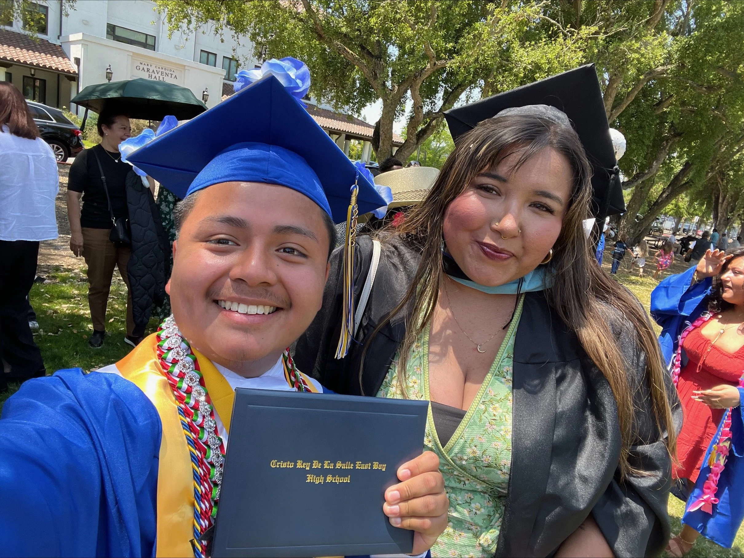 A young man and a woman celebrating a graduation outdoors, both wearing caps and gowns, holding a diploma, with trees and other people in the background.