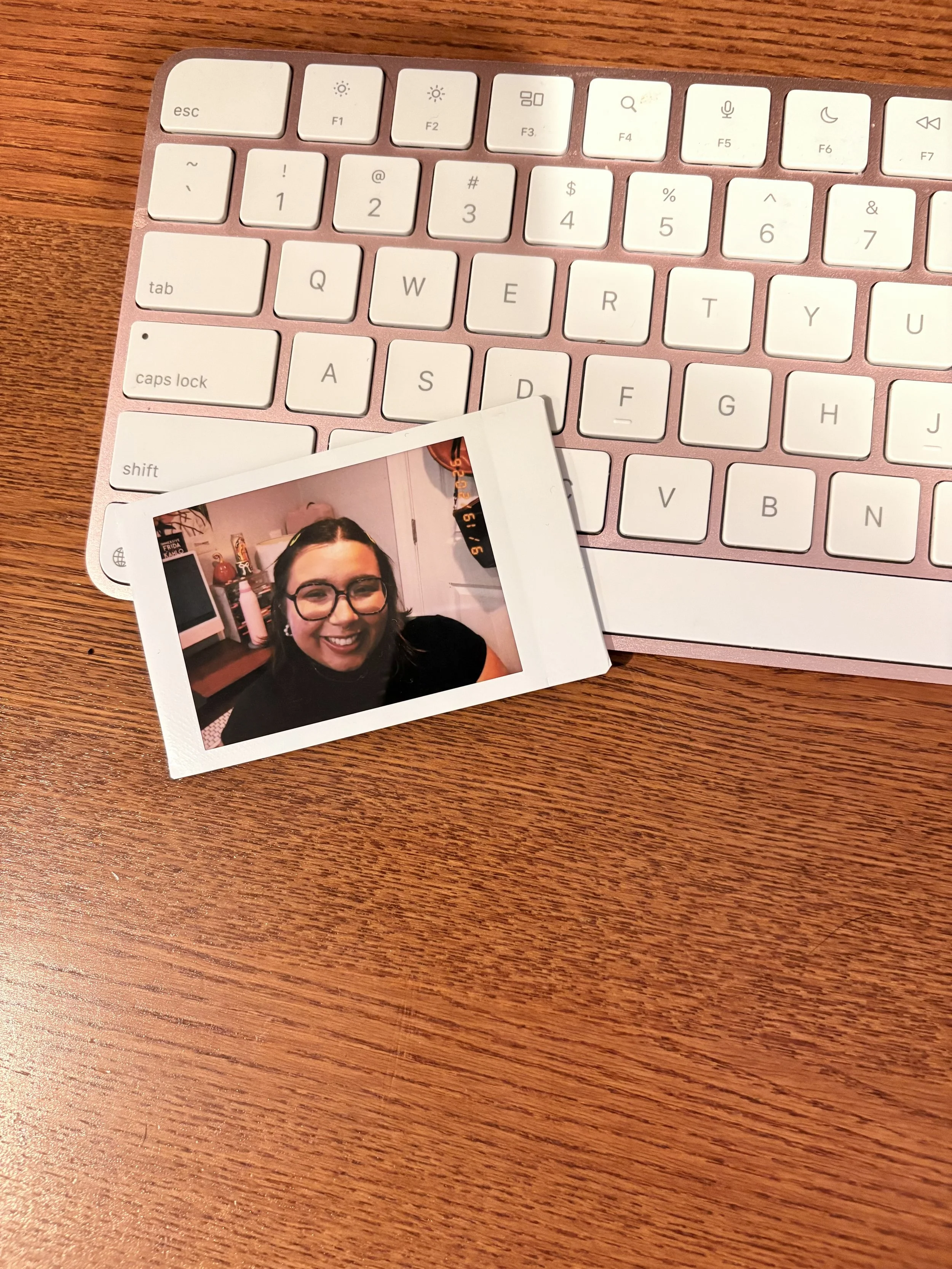 Close-up of a white computer keyboard with a pinkish metallic case on a wooden surface, with a printed photo of a smiling young woman with glasses placed on the keyboard.