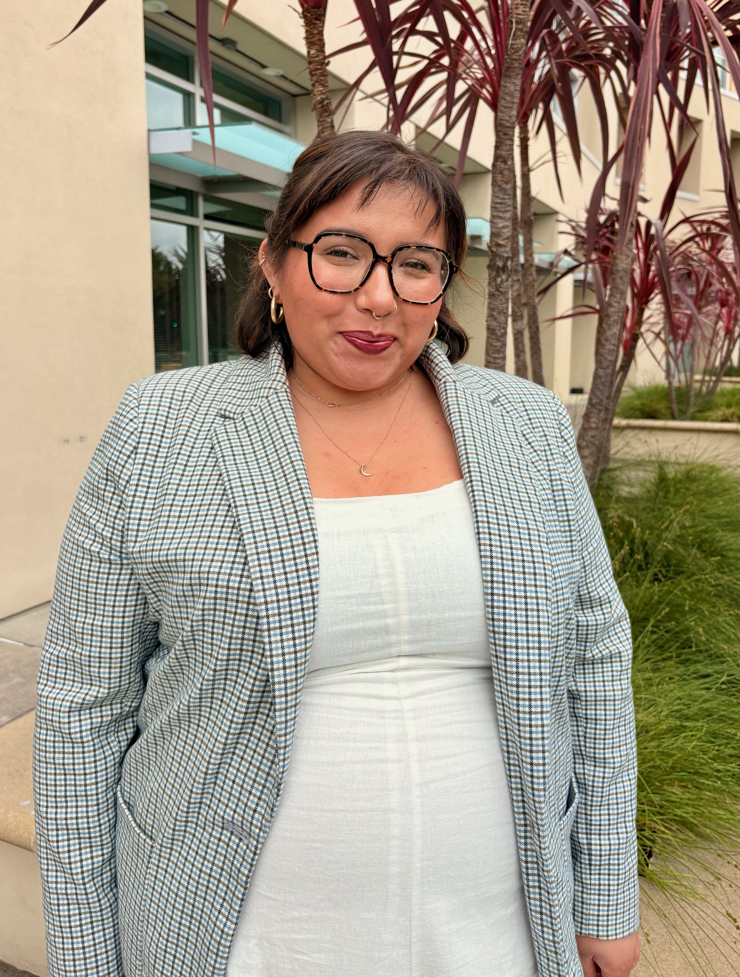 A woman with short brown hair, glasses, and earrings smiling outdoors in front of a tree and a building.