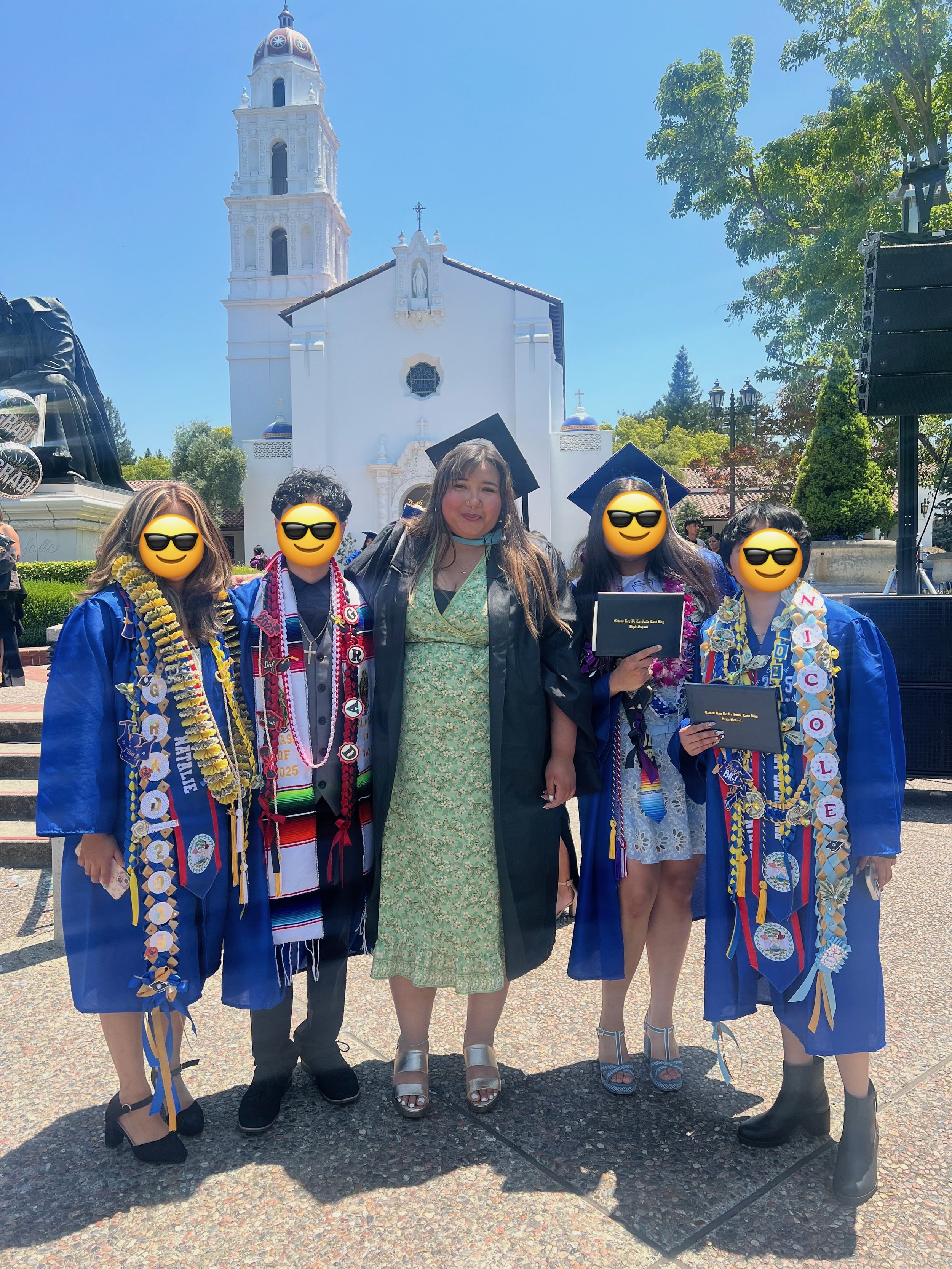 A group of five people in graduation caps and gowns standing in front of a white church, celebrating a graduation ceremony outdoors on a sunny day. The group appears joyful and proud.