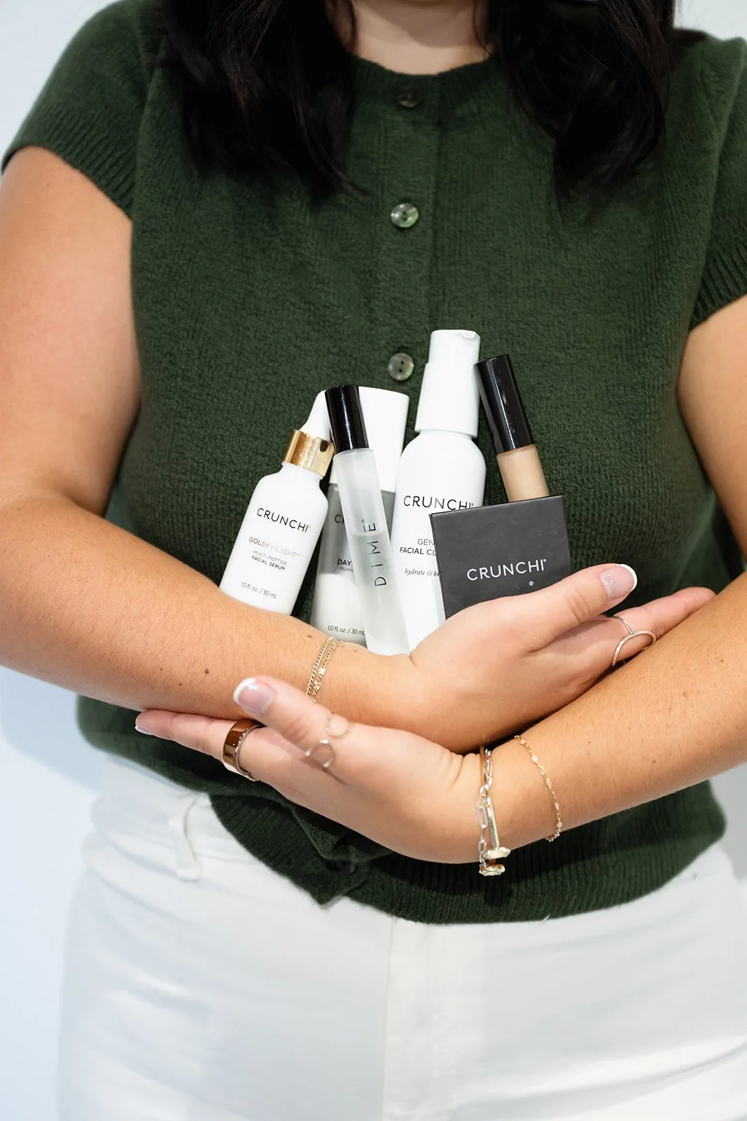 Person wearing a green shirt holding several skincare products, including CRUNCHI facial serums and sprays, against a plain background.