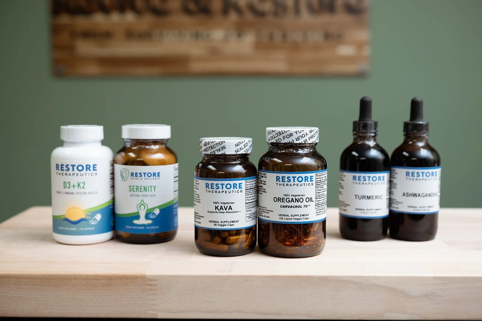 Bottles of herbal supplements from Restore Therapeutics, including D3+K2, Serenity, Kava, Oregano Oil, Turmeric, and Ashwagandha, placed on a wooden surface against a green wall.