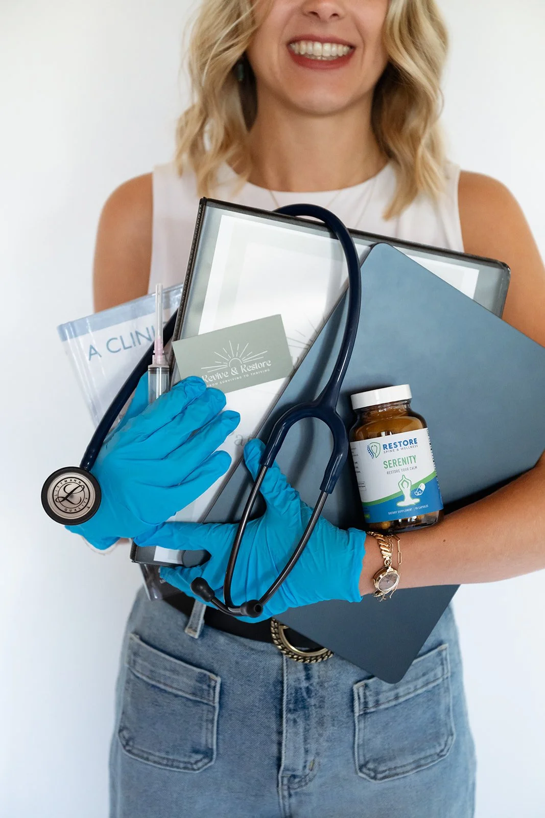 A woman holding a medical clipboard, stethoscope, medication, and office supplies, wearing blue gloves and a white sleeveless top.