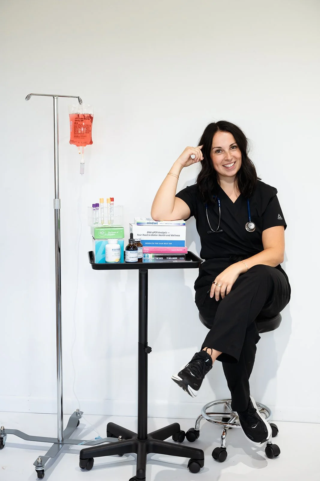 A healthcare professional sitting on a stool in a medical environment, smiling, with medical supplies and a IV stand nearby.