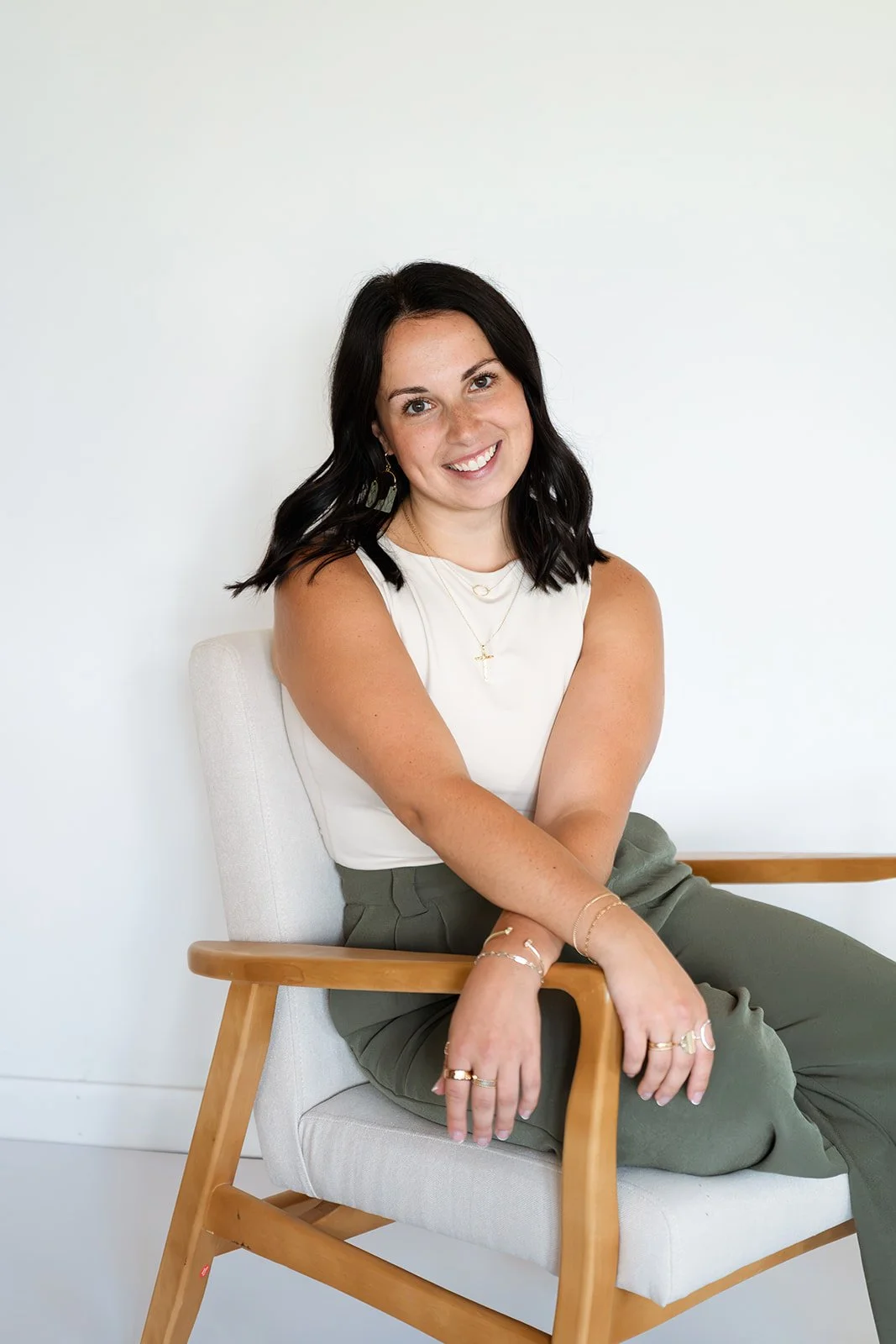 A woman with black hair sitting on a modern wooden chair with a light-colored cushion, smiling at the camera, wearing a white sleeveless top and green pants, against a plain white wall.
