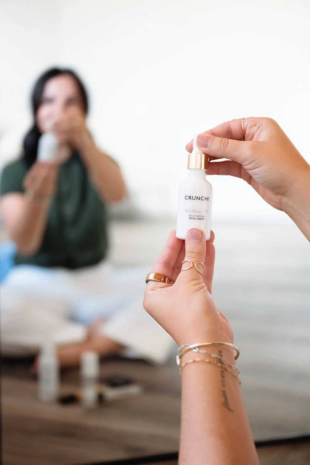 Close-up of hands holding a bottle of skincare serum labeled 'CRUNCHI' in focus, with an out-of-focus person in the background holding a mug, sitting on the floor in a bright room.