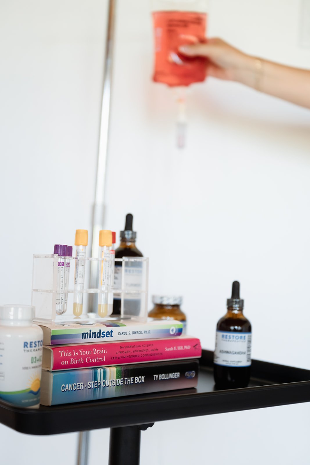 A black tray holding medical books, medicine bottles, and test tubes with yellow and purple caps. A hand is pouring a red liquid medicine from a bottle into a dropper above the tray.