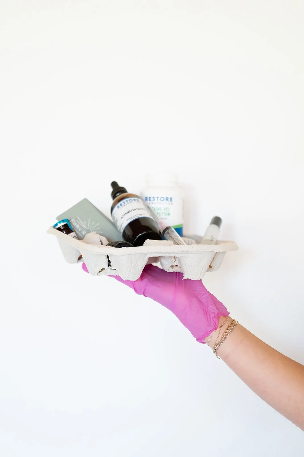 A hand wearing a pink glove holding a white paper tray with various medical supplies including bottles, syringes, and containers against a plain white background.