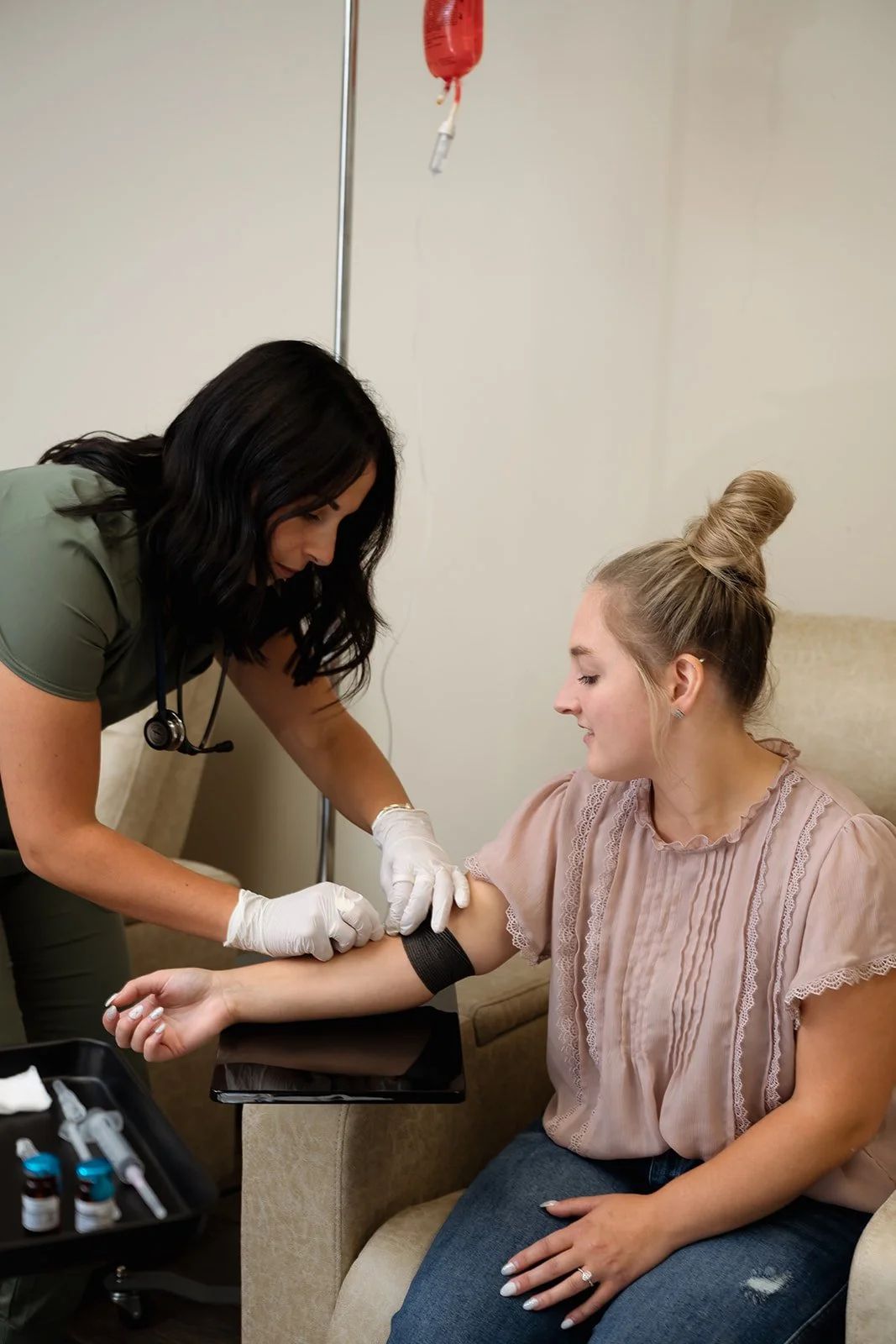 A nurse prepares to draw blood from a woman's arm in a medical setting, with a red IV bag hanging above them.