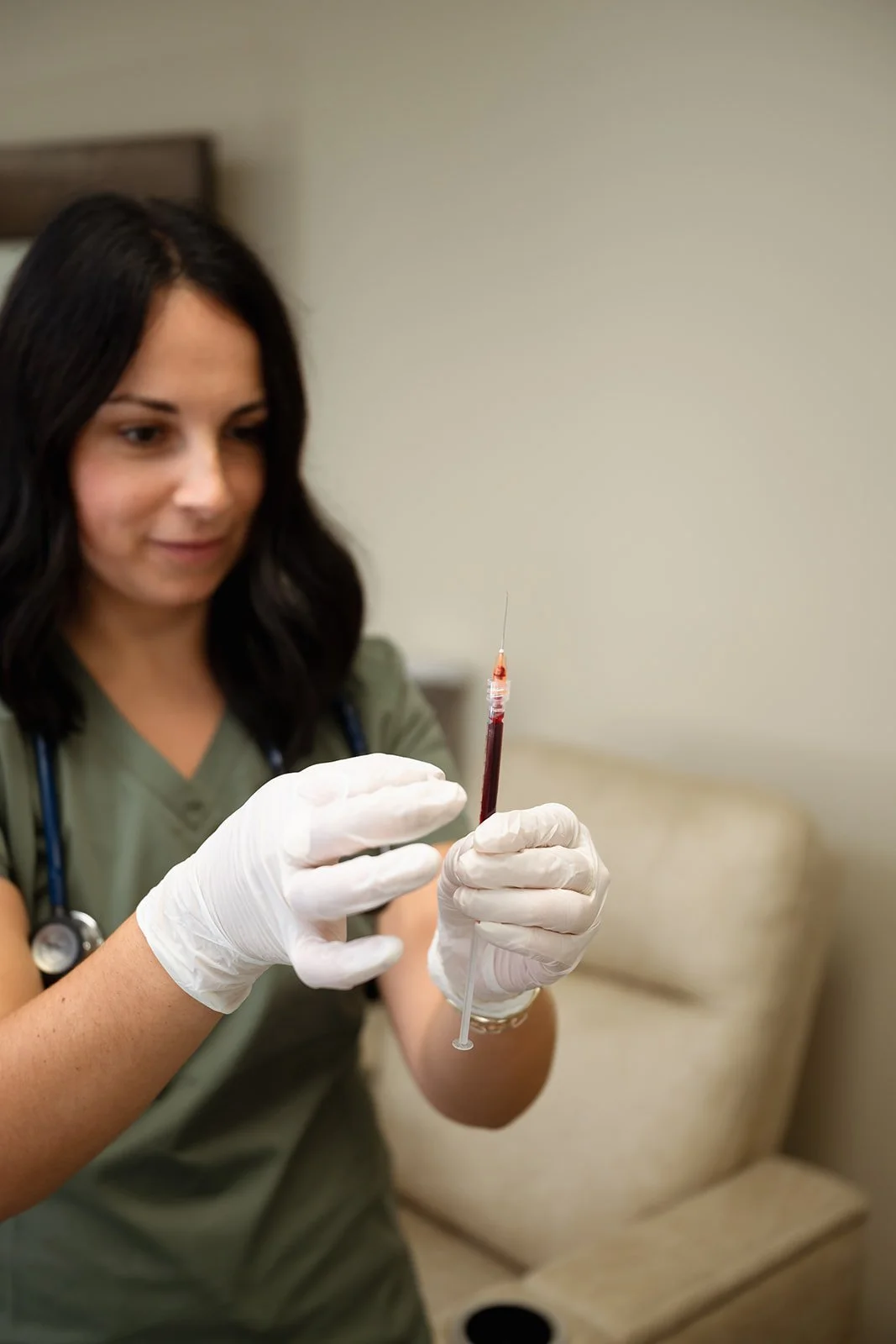 A female healthcare professional wearing gloves holds a syringe filled with blood, preparing a blood sample, with a stethoscope around her neck in a clinical setting.
