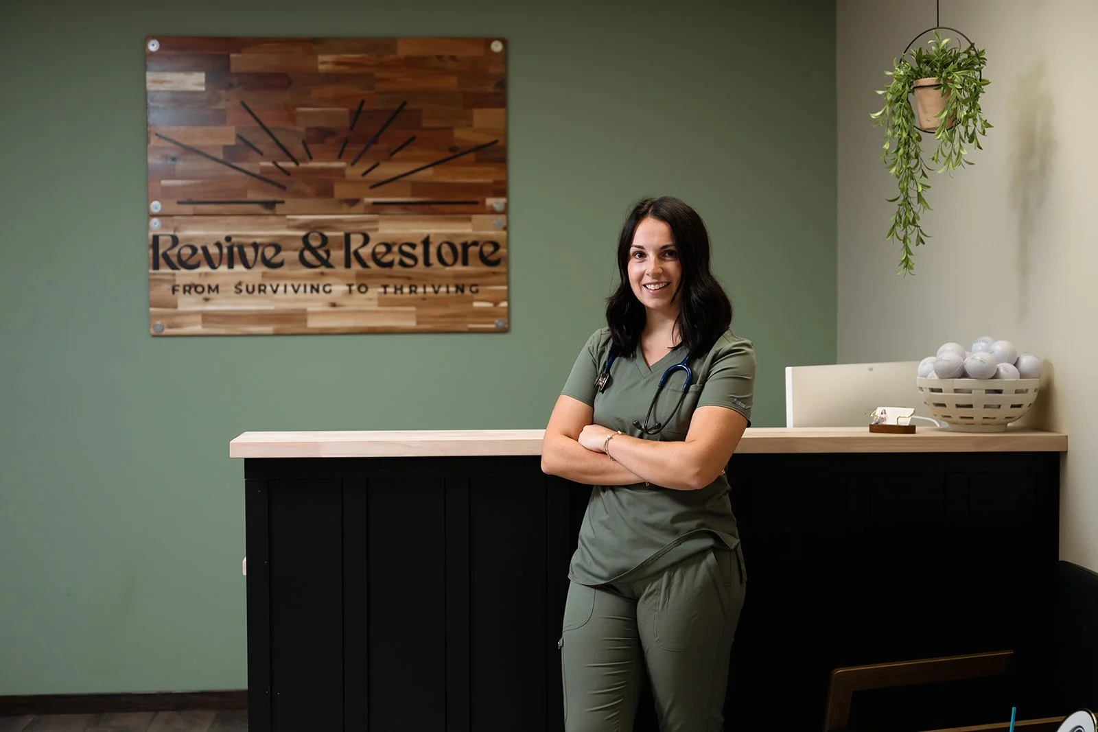 A woman in green scrubs with a stethoscope around her neck standing in a medical office. Behind her is a wooden reception desk with a basket of white balls and a hanging plant. A sign on the green wall reads 'Revive & Restore From Surviving to Thriving'.