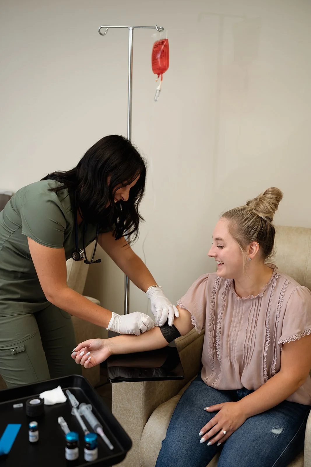 A nurse draws blood from a smiling woman in a medical clinic, with medical supplies and an IV stand in the background.