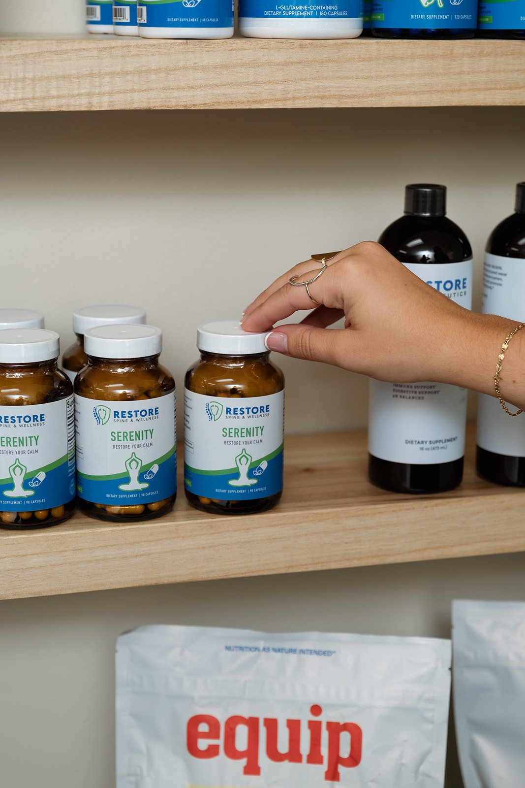 A person is reaching for a bottle labeled 'Restore Serenity' on a wooden shelf, with similar bottles and larger bottles behind. The bottles contain dietary supplements.