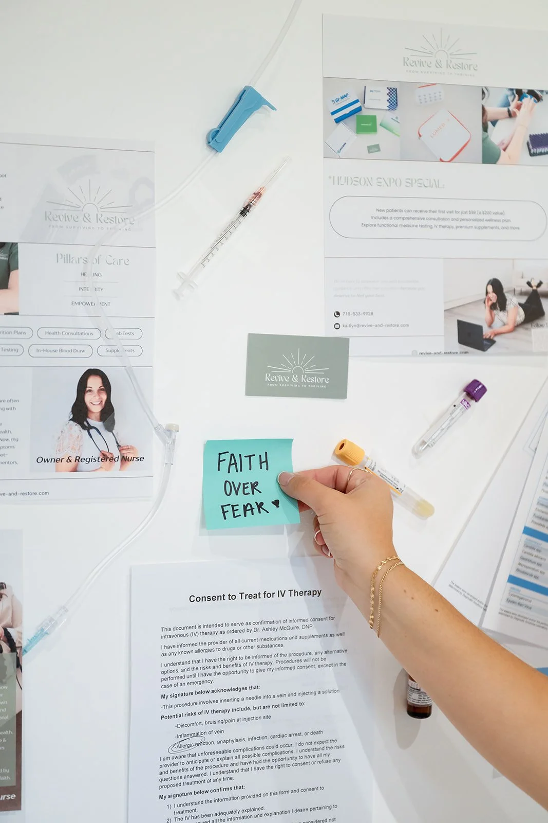 A desk with medical documents, syringes, and a person holding a sticky note that says 'FAITH OVER FEAR' amid healthcare items.