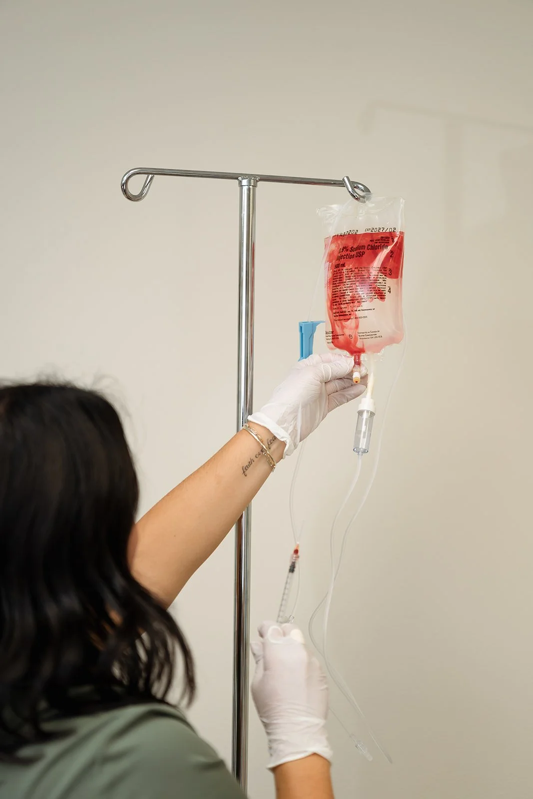 A healthcare professional prepares an intravenous medication bag, hanging on a metal stand, with a syringe and tubing, in a medical setting.