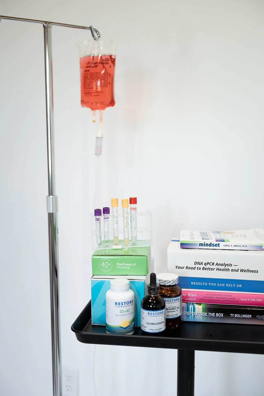 An IV drip bag hanging on a stand with a fluid infusion, and a tray with test tubes, medication bottles, and medical books on health and wellness.