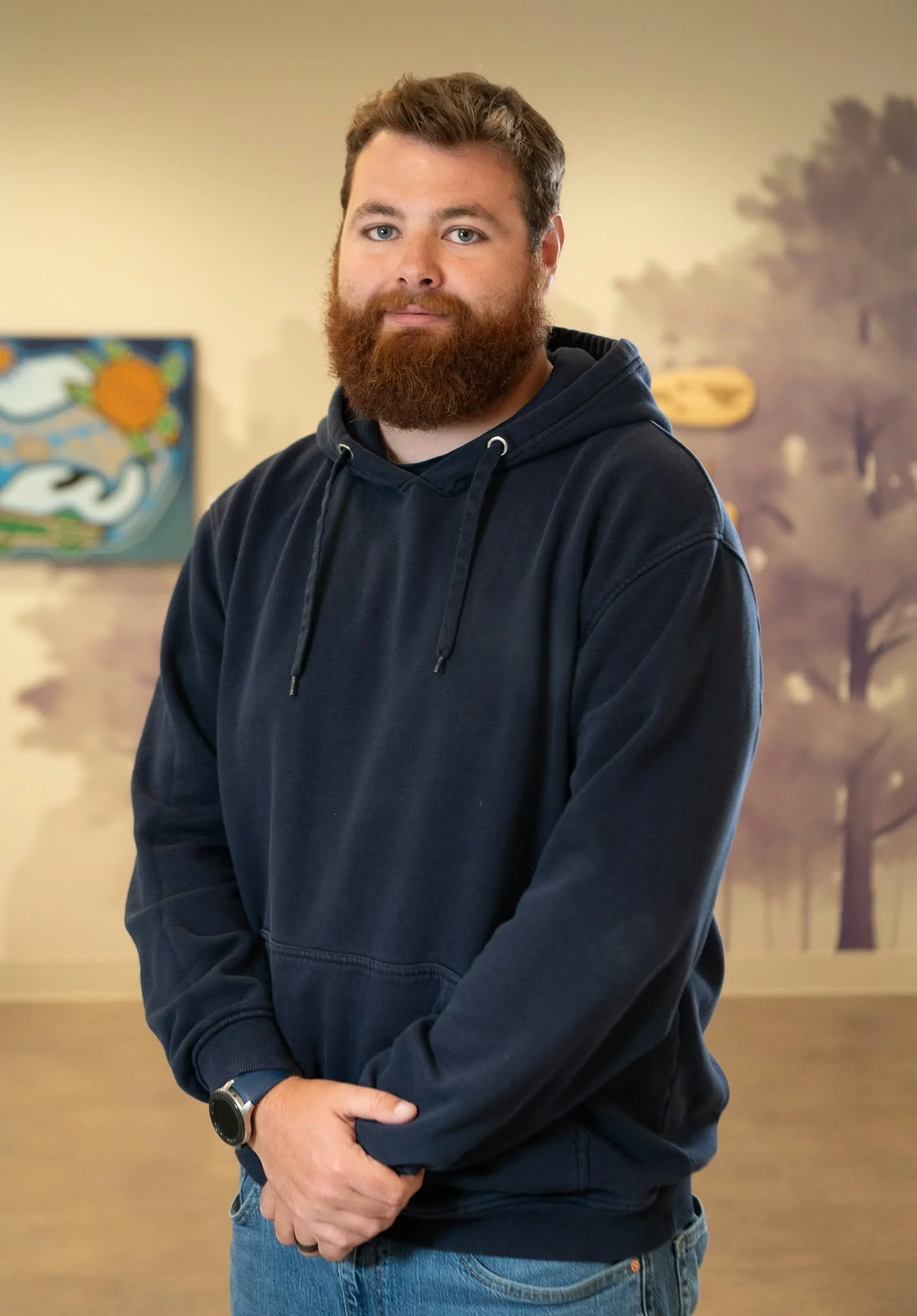 A man with a beard and blue eyes wearing a black hoodie and blue jeans standing in a room with a blurred background of artwork and trees.