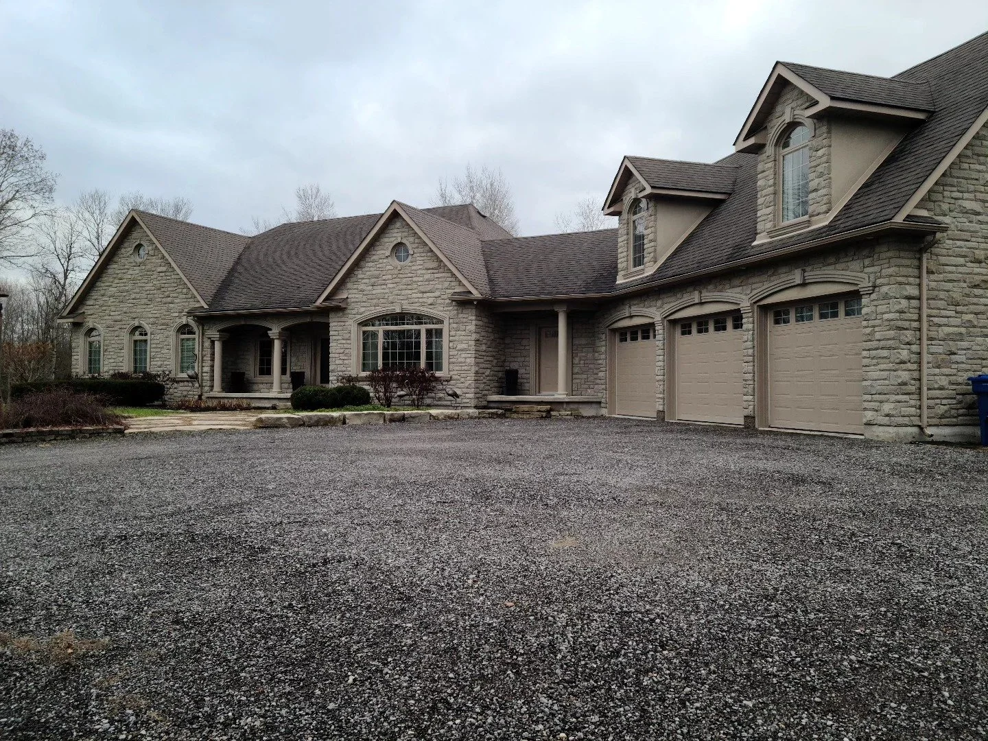 Large stone house with a gravel driveway and attached three-car garage, surrounded by leafless trees on overcast day.