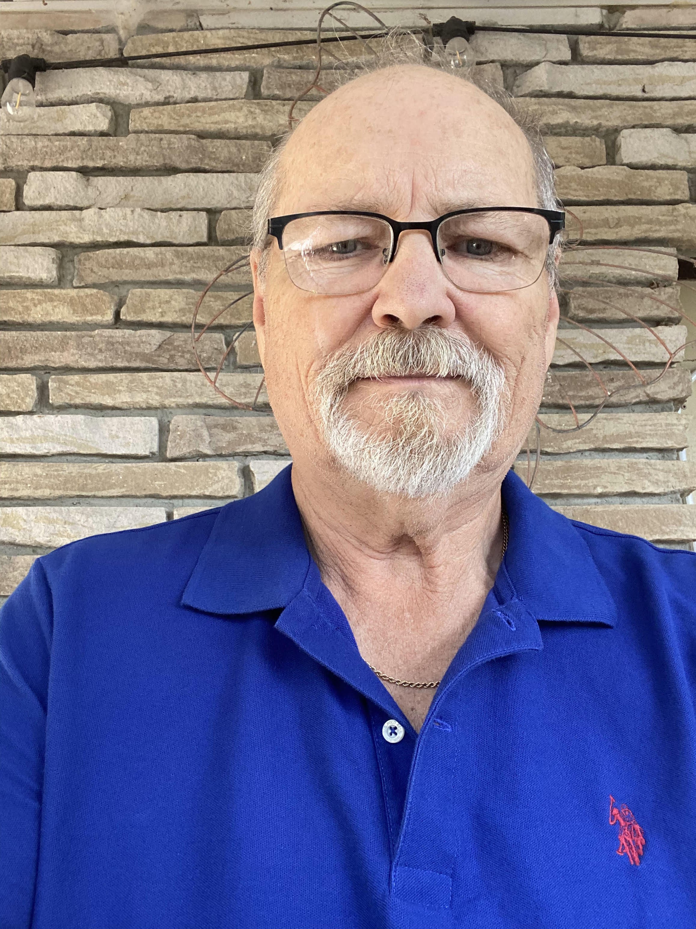 Close-up of an older man with a beard and glasses, wearing a blue polo shirt with a red embroidered logo, standing in front of a brick wall with exposed wiring and lights.