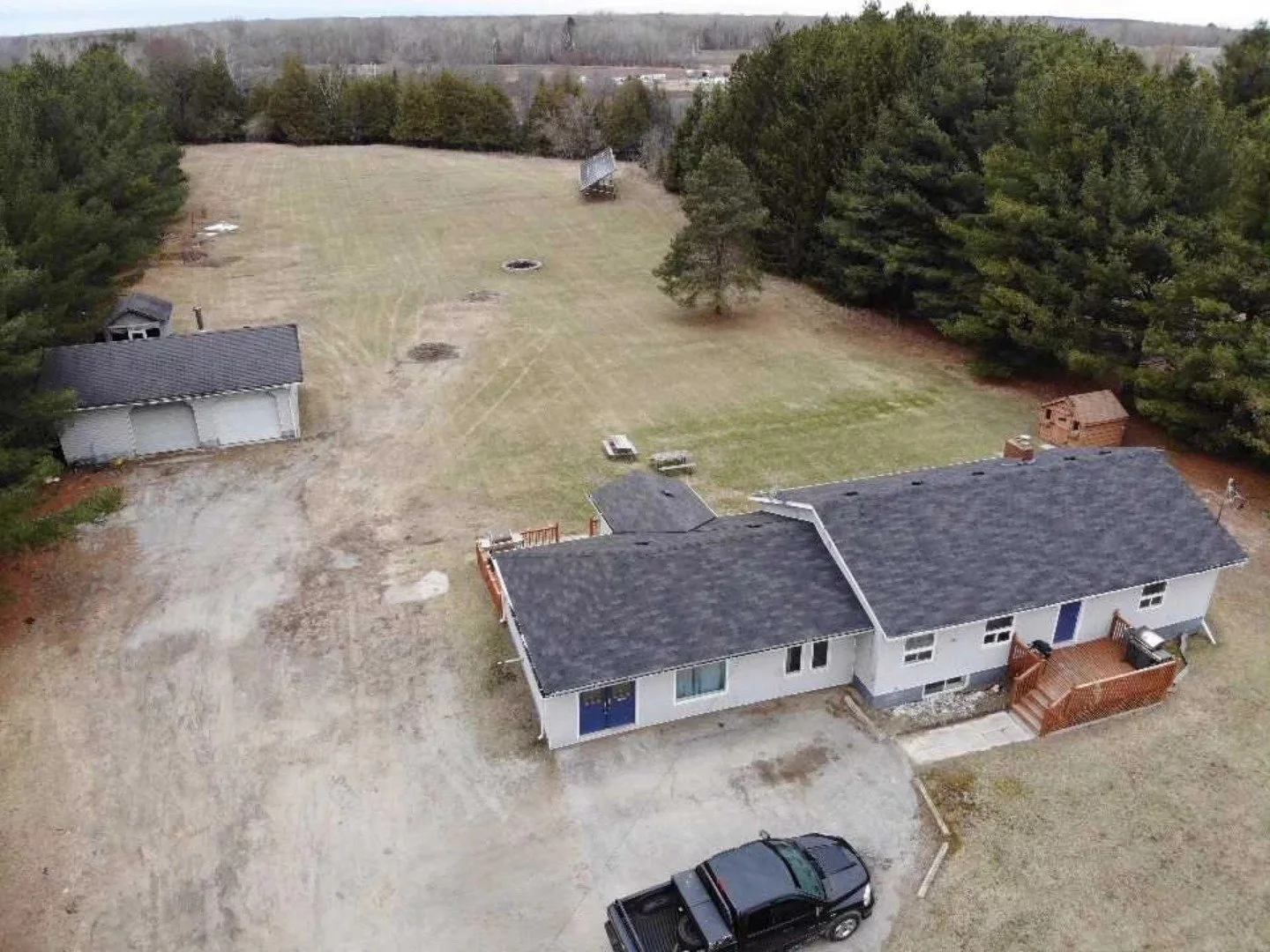 Aerial view of a house with a black roof, a parking area with a black vehicle, and a large backyard with scattered trees, sheds, and an open grassy field with some patches of dirt.