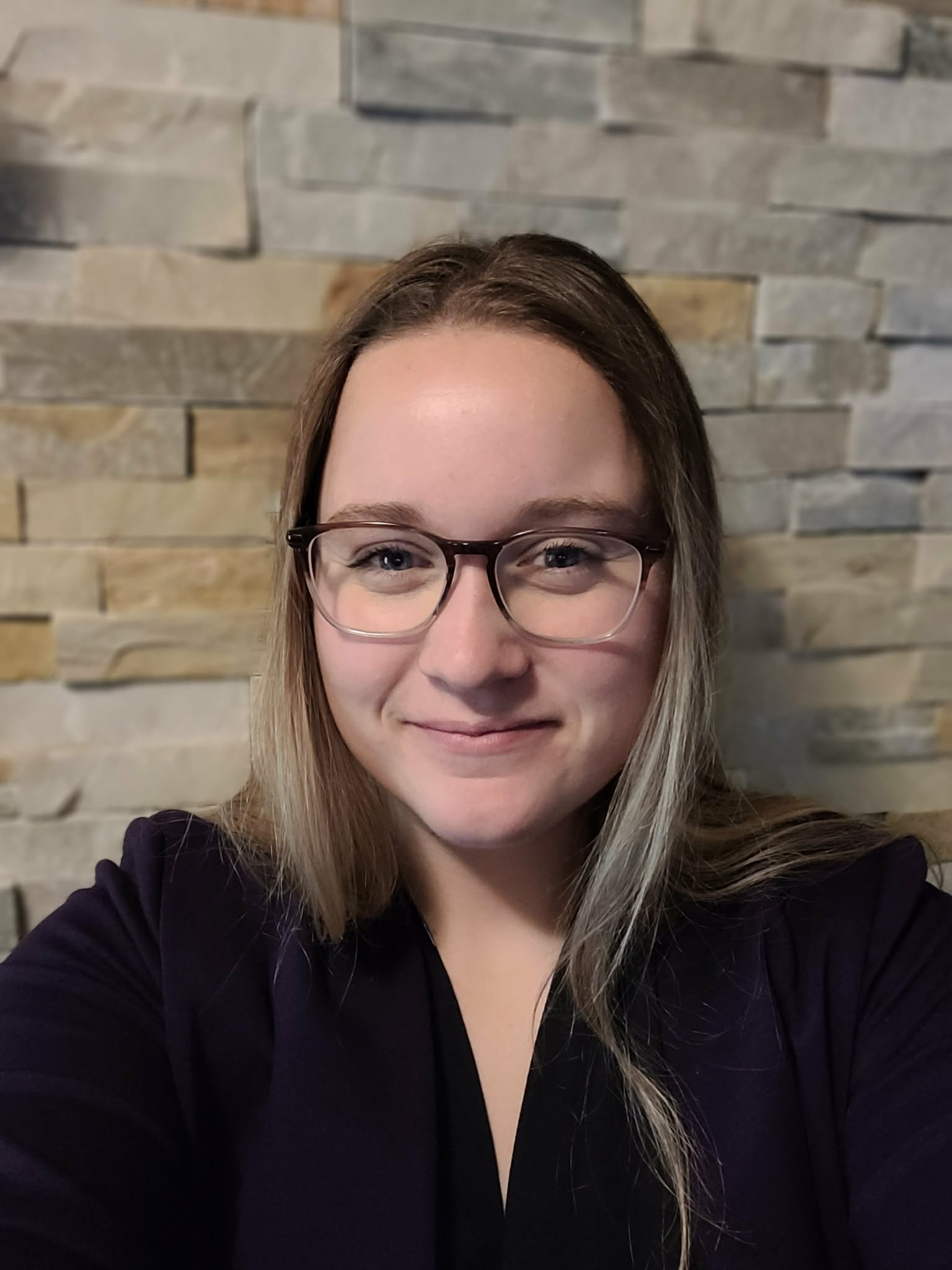 A young woman with blonde hair, glasses, and a black top smiling in front of a stone wall background.