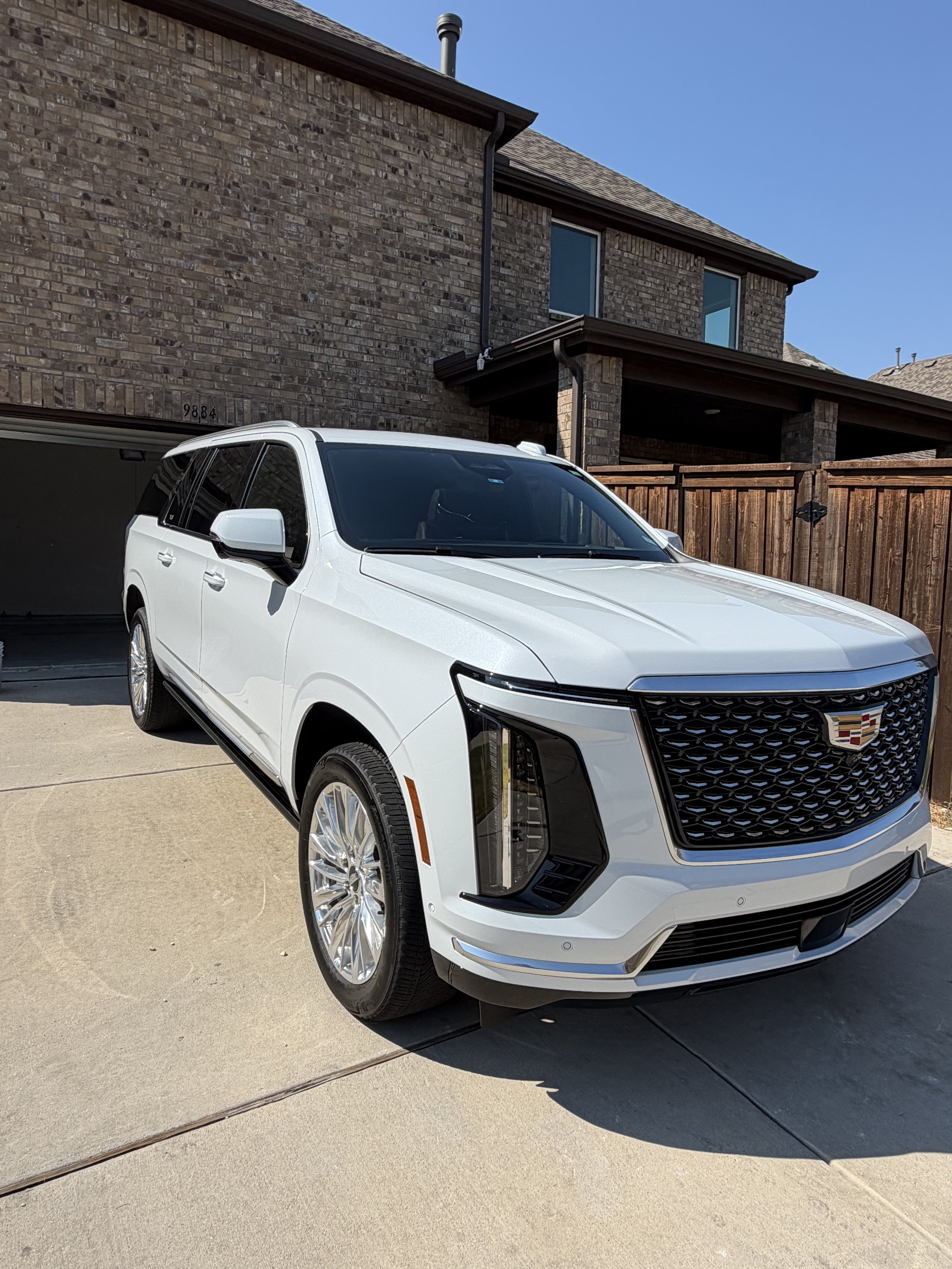 Front view of a white Cadillac SUV parked on a driveway in front of a brick house with a wooden fence, under a clear blue sky.