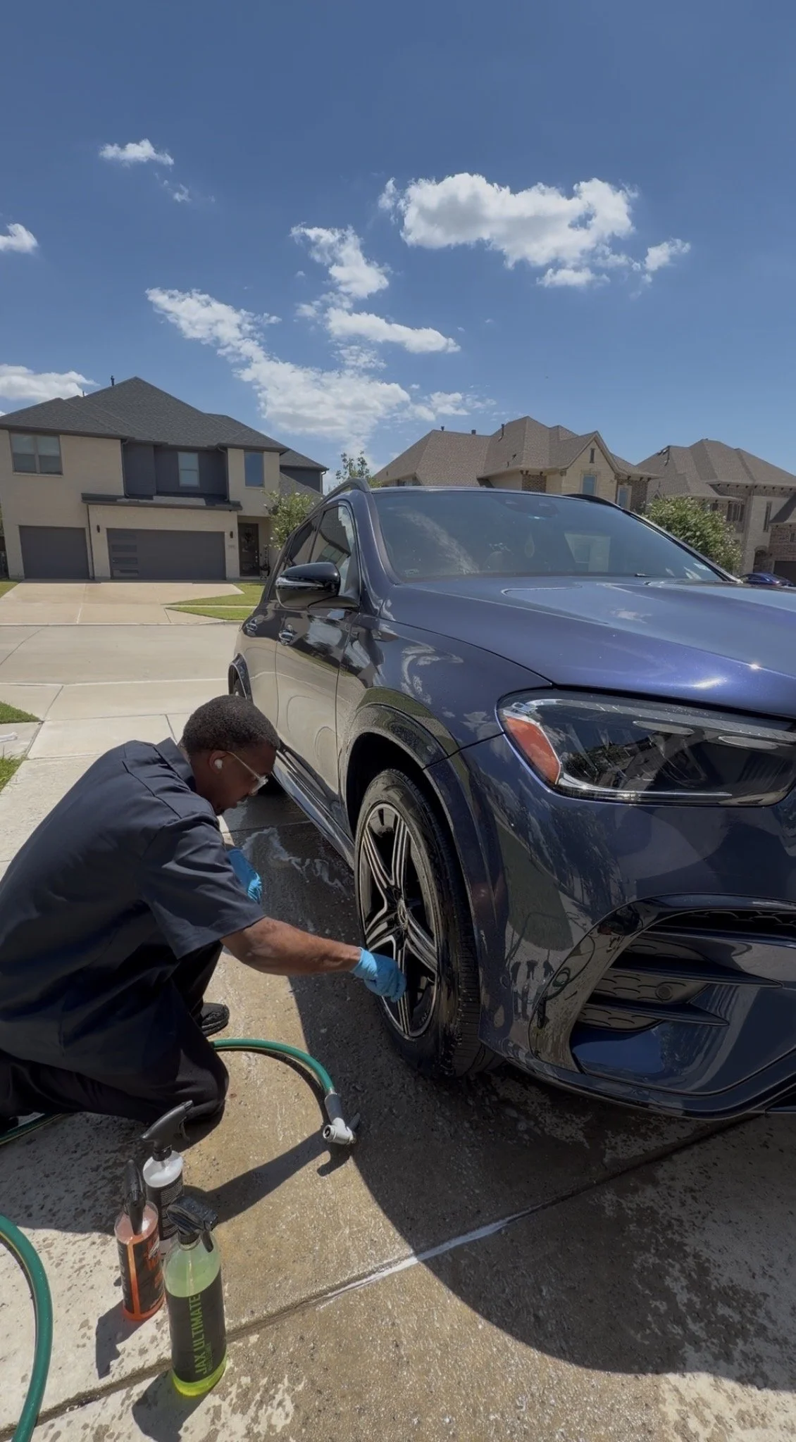 A man is washing a black SUV on a driveway in a residential neighborhood with multiple houses and a blue sky with clouds overhead.